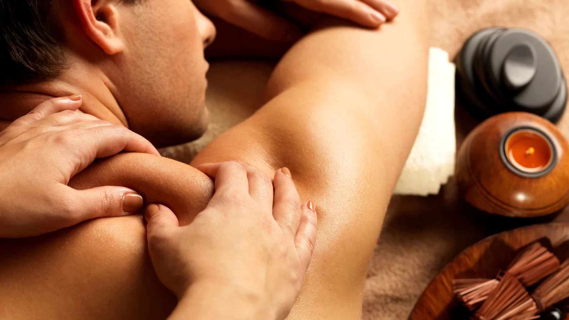 Man receiving shoulder massage with spa candle and warm therapy tools beside treatment table