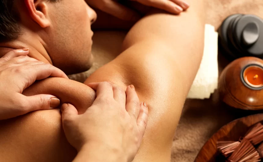 Man receiving shoulder massage with spa candle and warm therapy tools beside treatment table