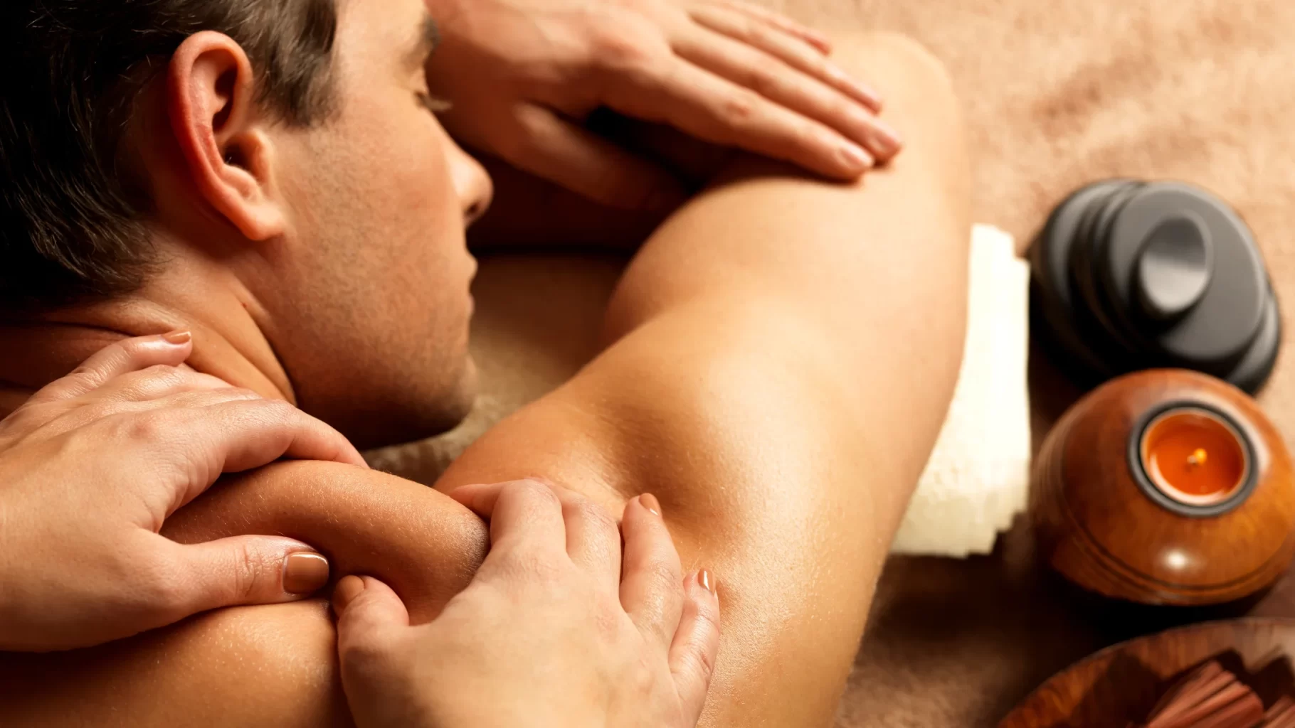 Man receiving shoulder and upper back massage with spa candle and warm therapy tools nearby