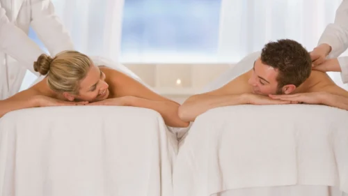 Couple receiving side-by-side massage treatments on spa tables in a bright and peaceful wellness room.