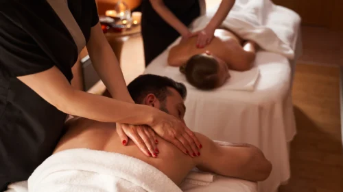 Couple receives relaxing back massages from spa therapists in a warm, softly lit spa treatment room.