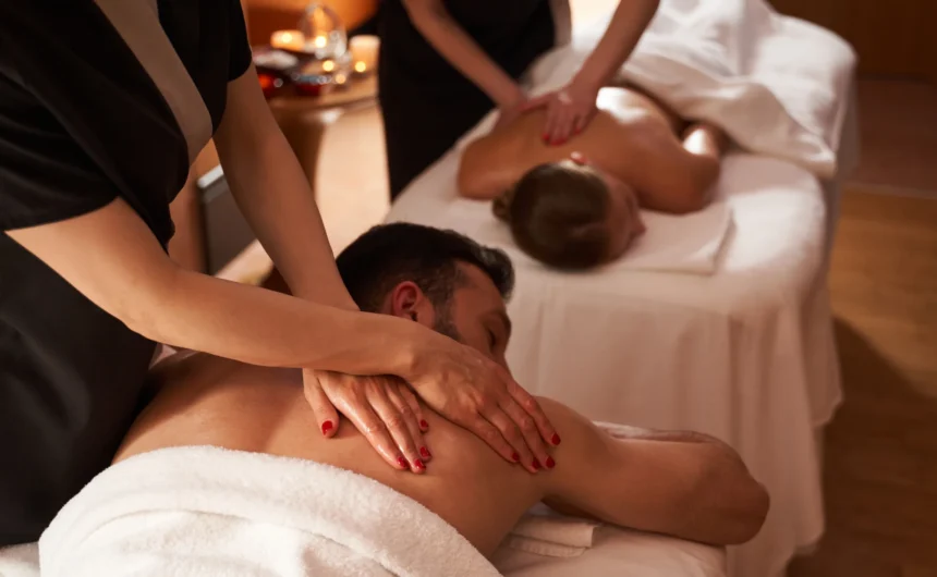 Couple receives relaxing back massages from spa therapists in a warm, softly lit spa treatment room.