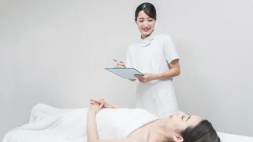 Spa professional checks a clipboard beside a client resting on a treatment bed before a spa service.