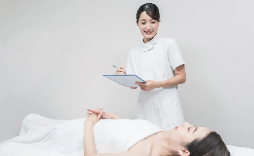 Spa professional checks a clipboard beside a client resting on a treatment bed before a spa service.